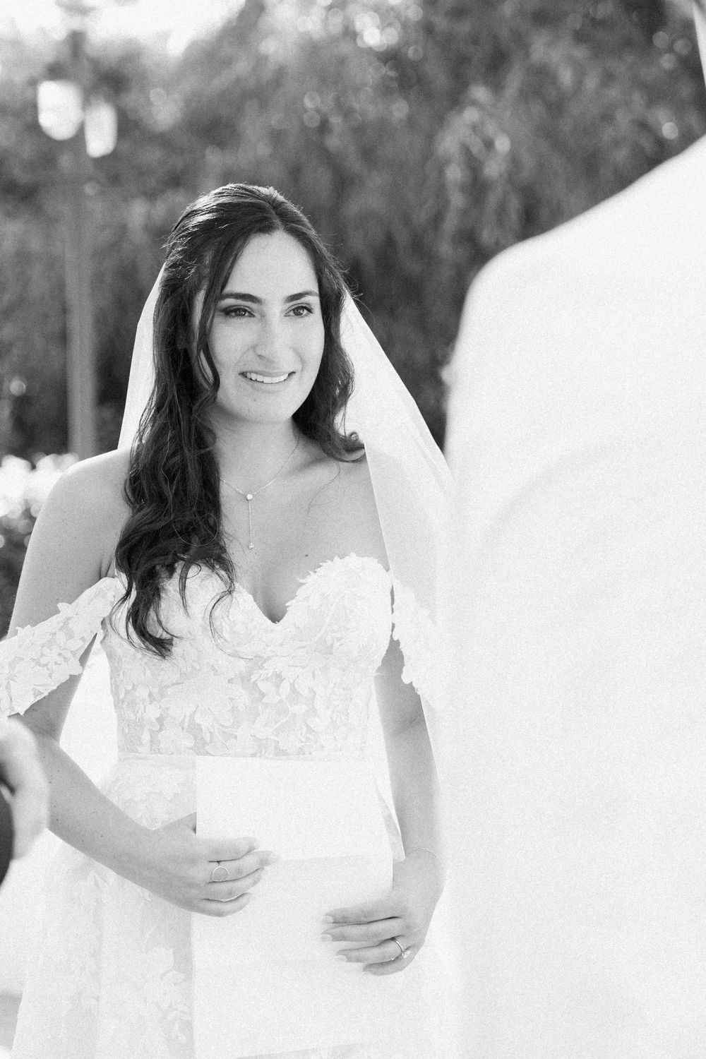 Black and white photo of a smiling bride in lace off-shoulder gown and veil during outdoor wedding ceremony.