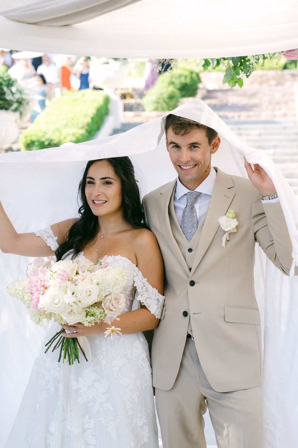 Bride and groom smiling under flowing white veil, bride holding pink floral bouquet, groom in beige suit at outdoor wedding.