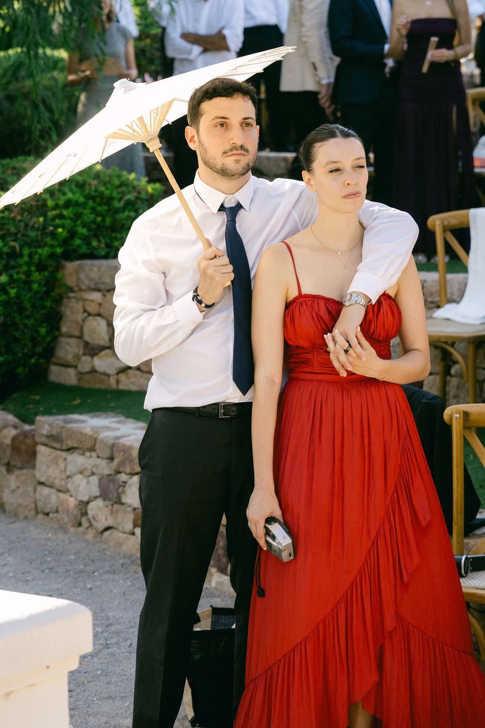 Elegant couple at outdoor event, man in white shirt holding parasol, woman in red gown, garden setting.