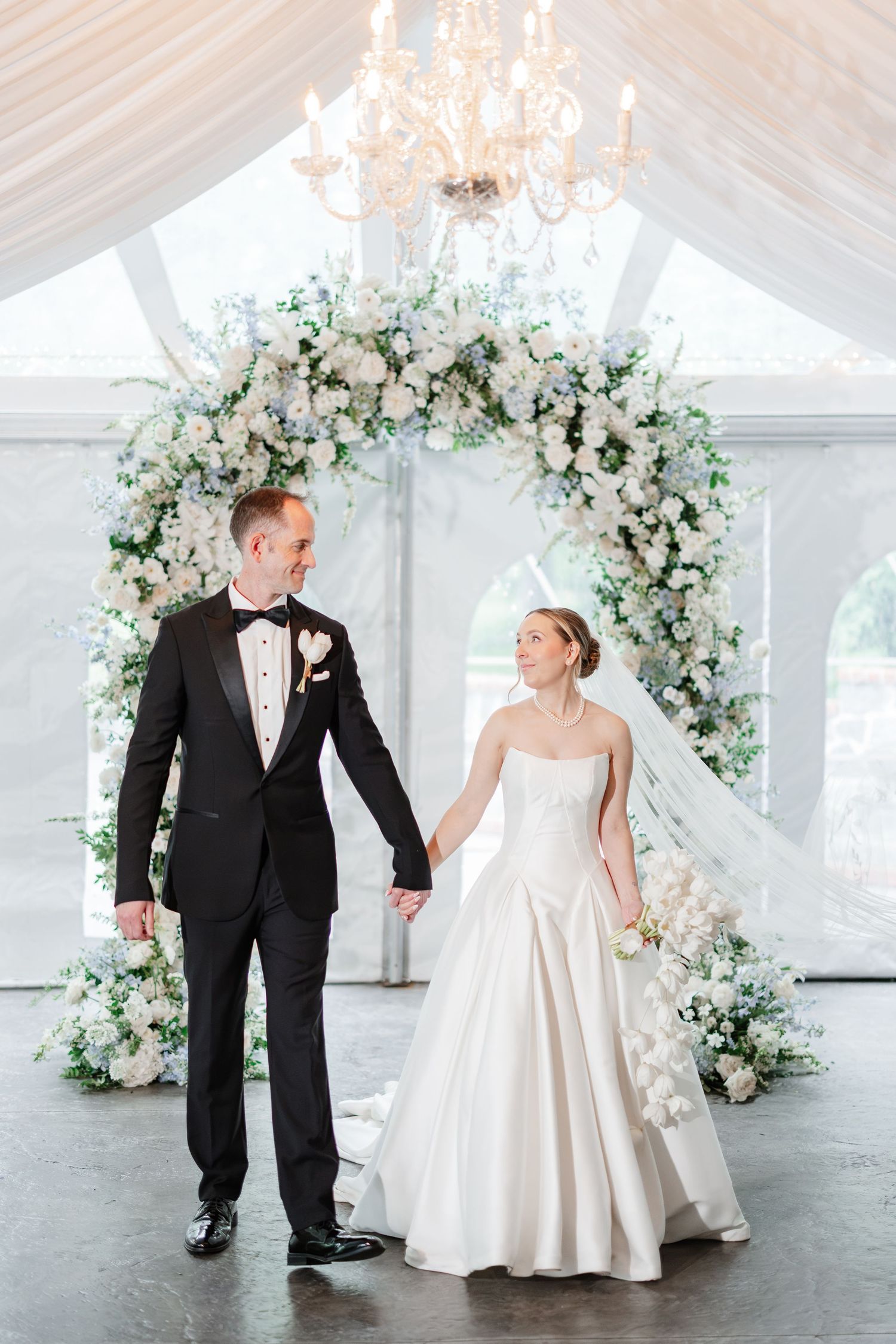 Bride and groom holding hands under floral arch of white and blue blooms in elegant tented venue.