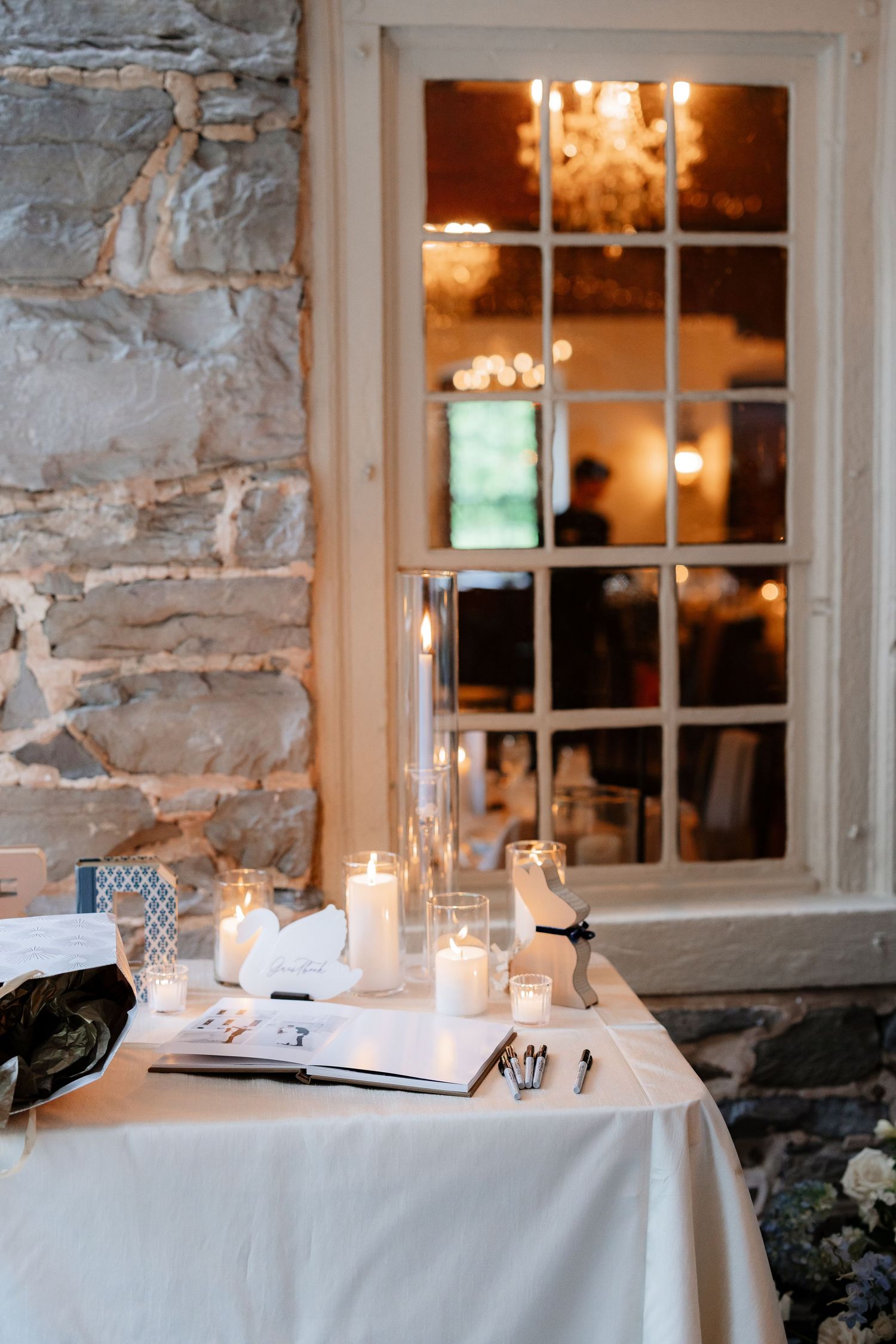 Wedding guest book table with candles beside stone wall and window reflecting warm chandelier light.