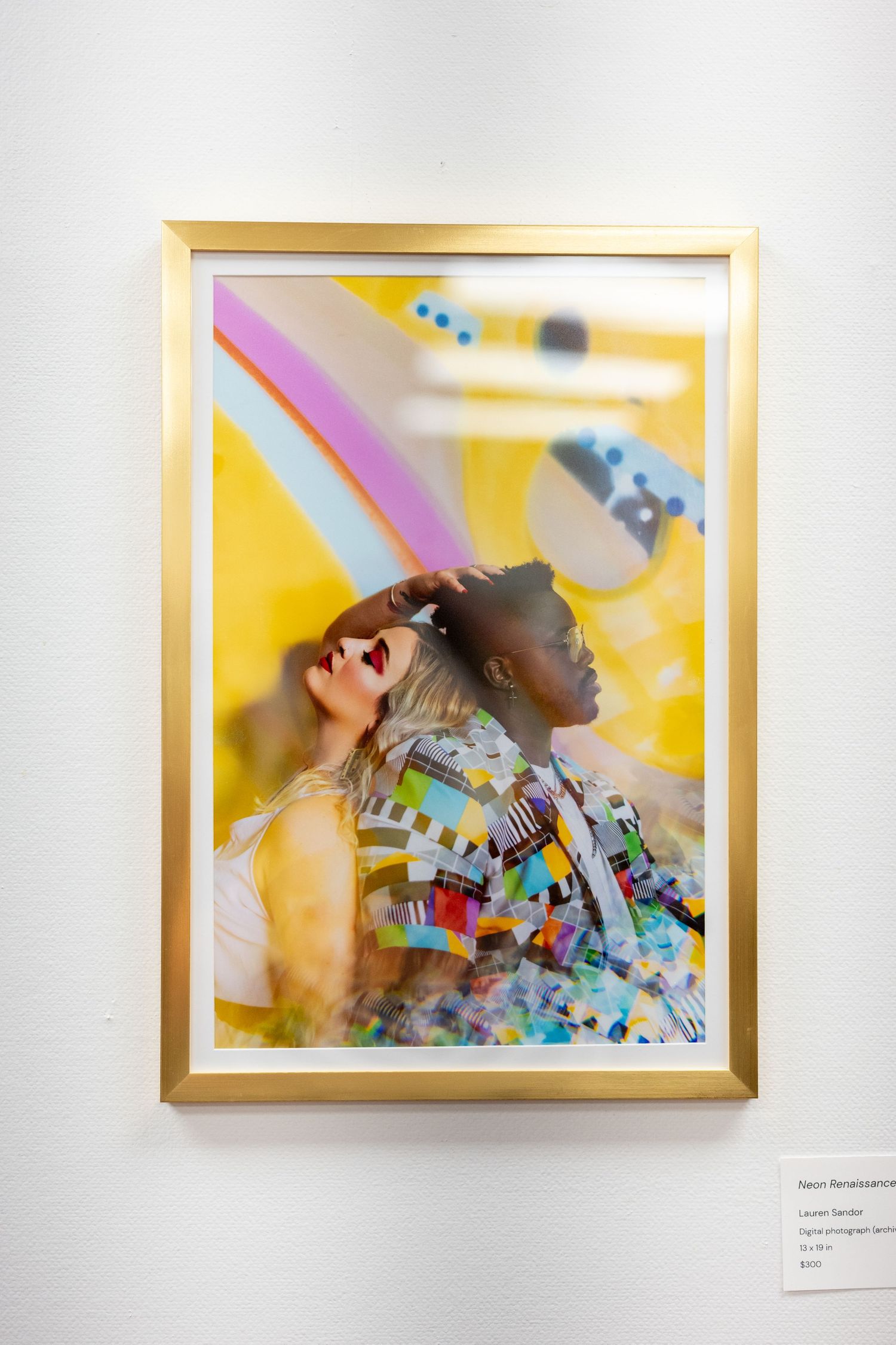 Gold-framed art photo of two people in colorful attire against a vibrant yellow background with rainbow elements.