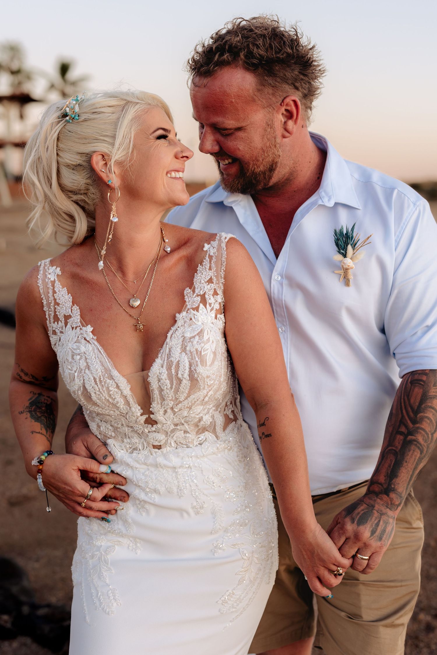 Couple in wedding attire holding hands on beach, bride in lace gown, groom in white shirt with boutonniere.