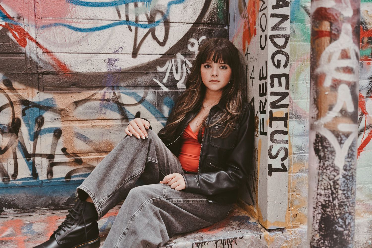 Young woman in leather jacket and grey jeans sits against graffiti wall with urban street art backdrop.