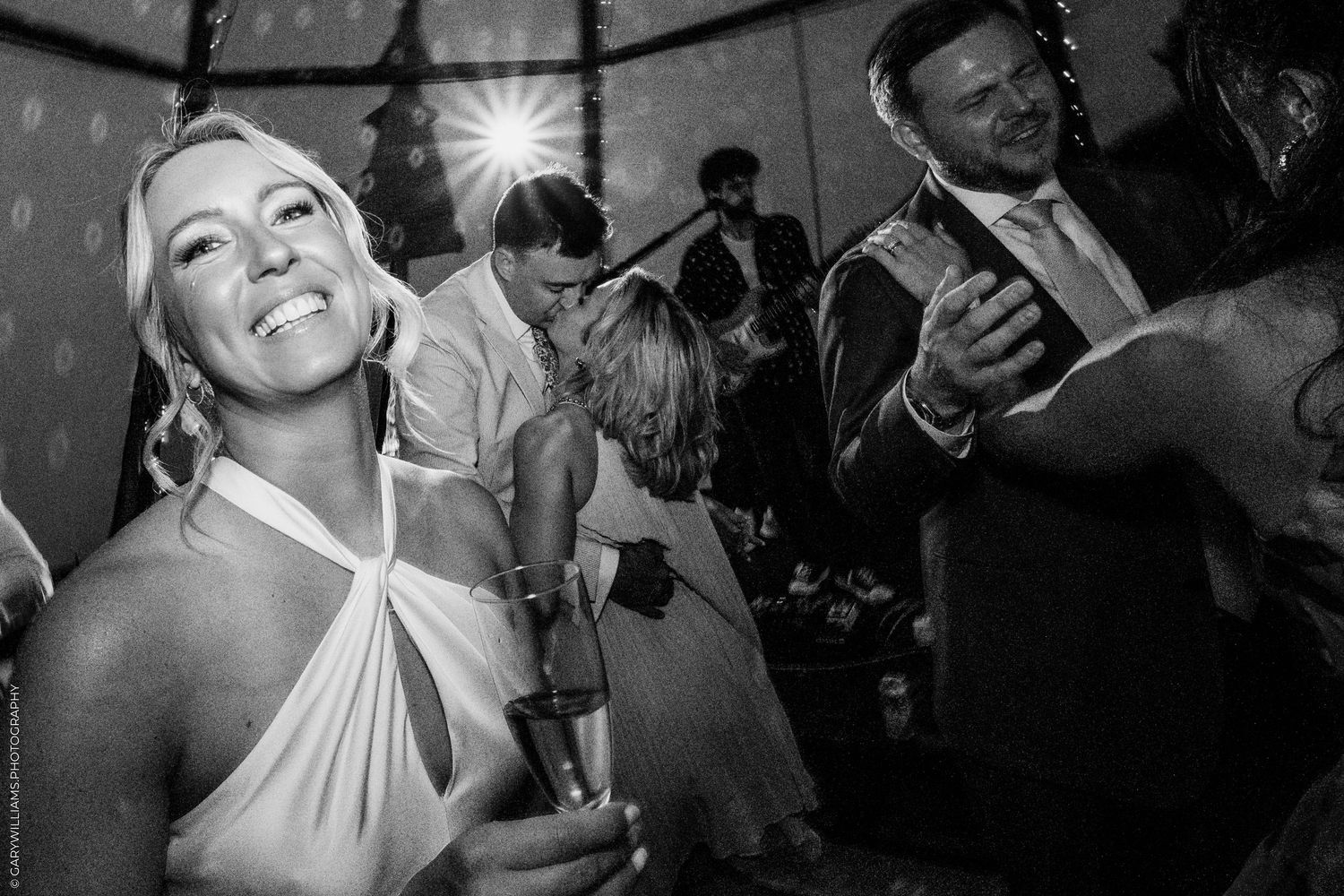 Black and white photo of a smiling woman in a white halter dress holding champagne at a lively wedding reception dance floor.
