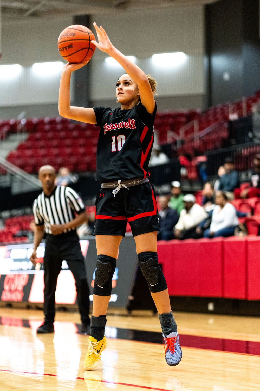 A female basketball player in black and red uniform shoots a jump shot during a high school game.