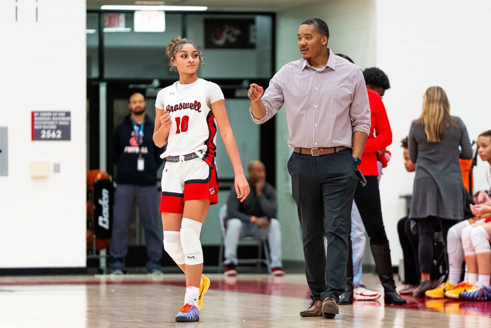 A female basketball player in white uniform walks alongside a coach during a high school basketball game.