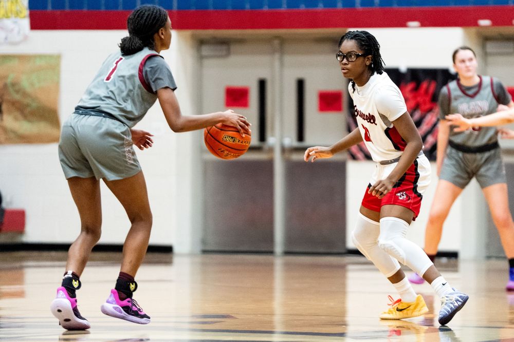 Two female basketball players compete on defense during a high school basketball game in a red gymnasium.