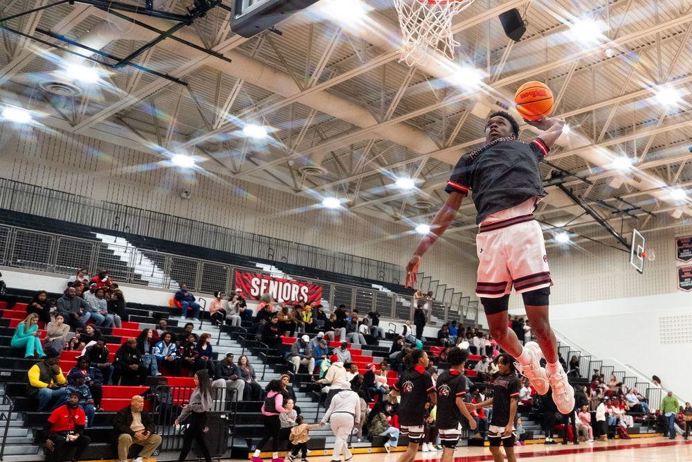 A basketball player soars for a spectacular dunk in front of a packed gymnasium crowd during a high school game.