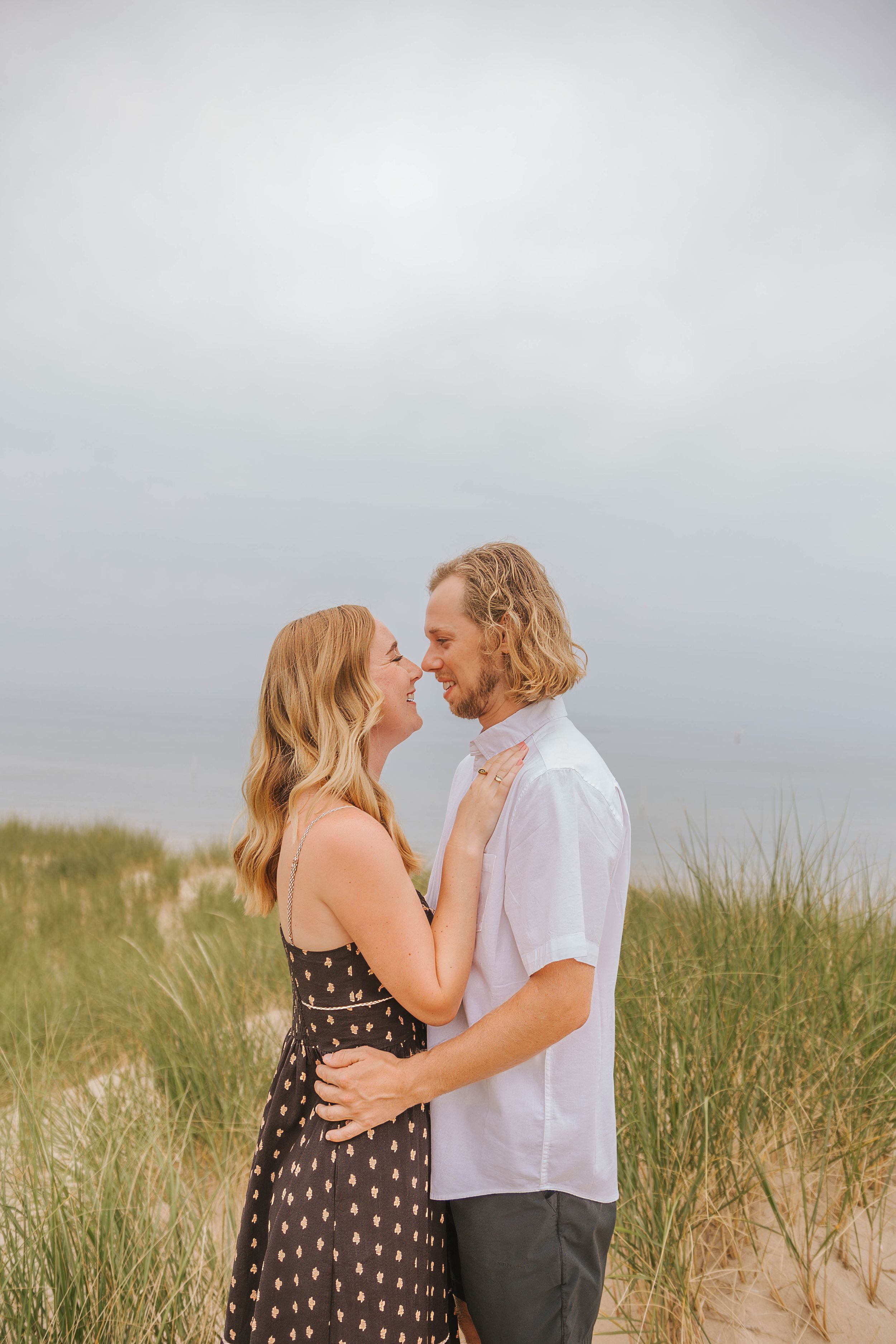 Couple embracing and smiling at each other on sandy beach dunes with ocean backdrop on overcast day.