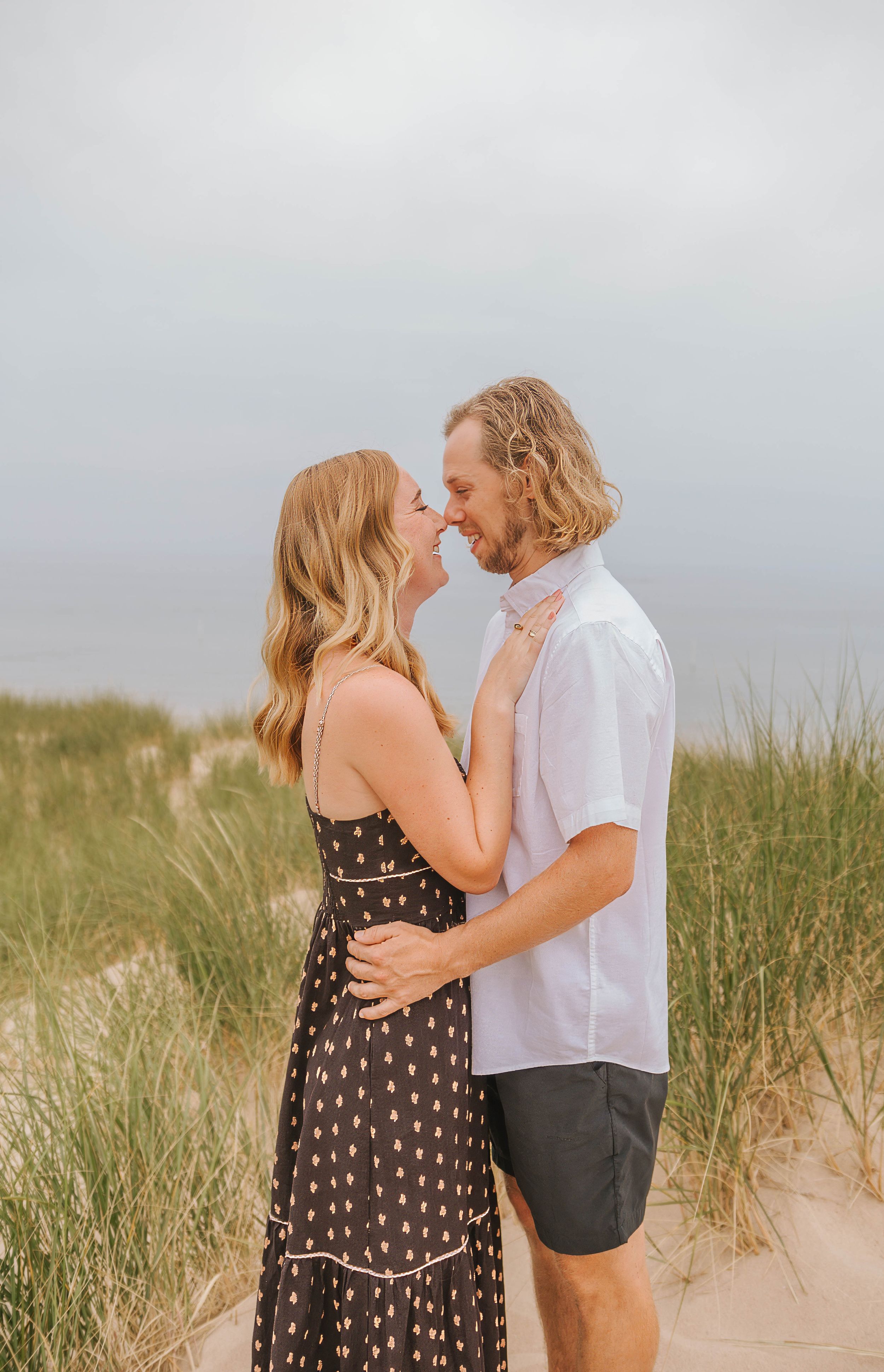 Couple sharing a romantic almost-kiss on sandy beach dunes, woman in floral dress, man in white shirt and dark shorts.