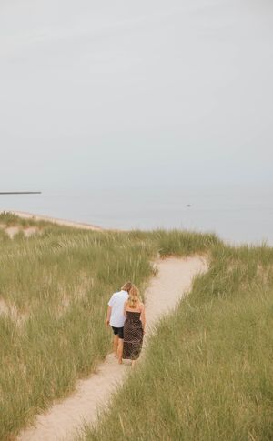 Couple walking hand in hand on a sandy beach path through green dune grass toward the ocean.
