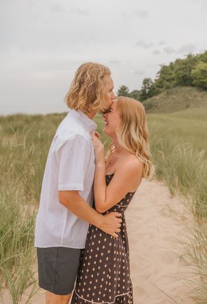 A young couple shares a romantic moment on a sandy beach path surrounded by dune grass on a cloudy day.