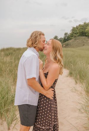 A couple shares a romantic moment on a sandy beach path surrounded by dune grass, the woman in a dark floral dress.