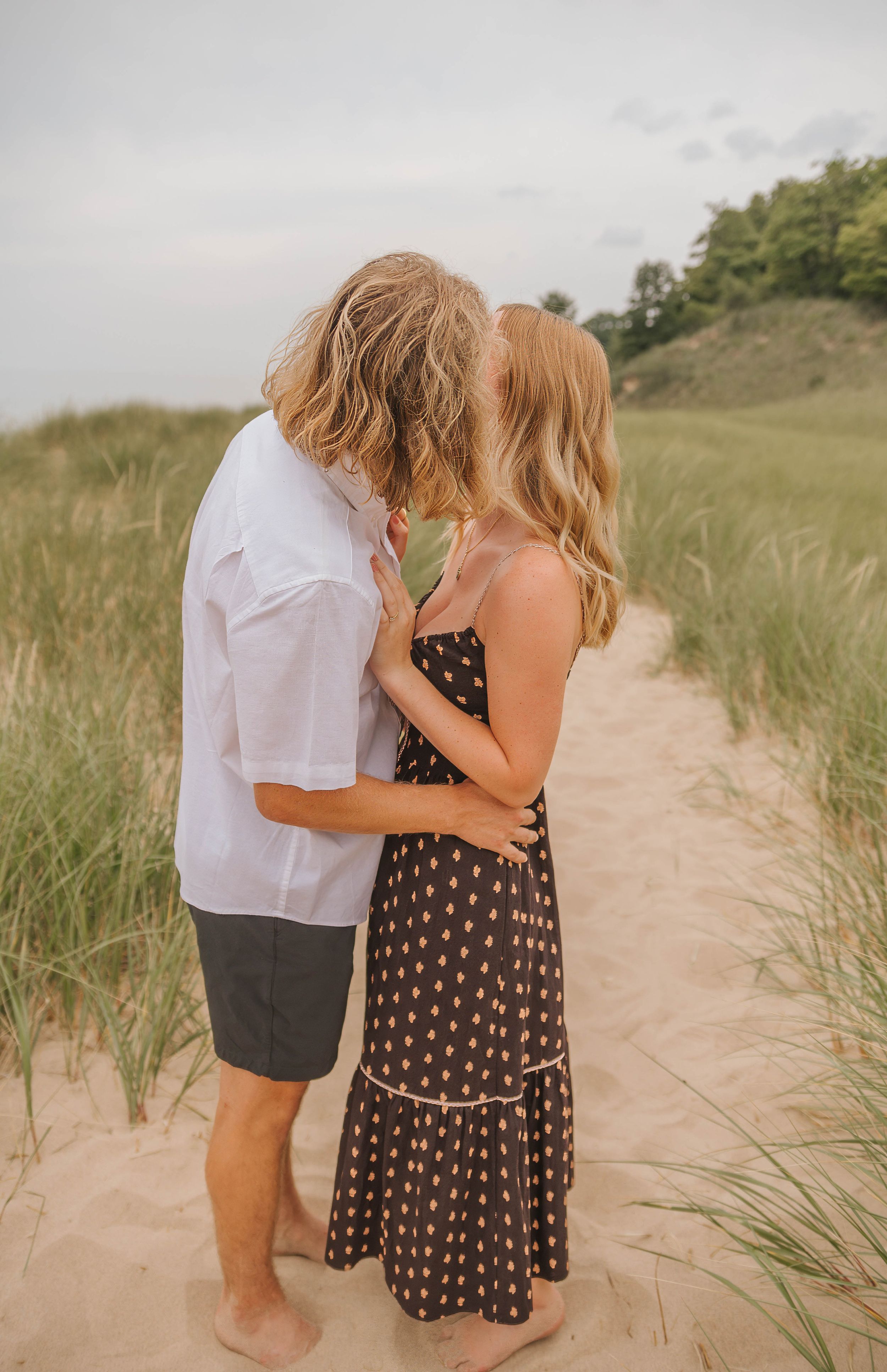 A couple shares a romantic kiss on a sandy beach path surrounded by dune grass on a cloudy day.