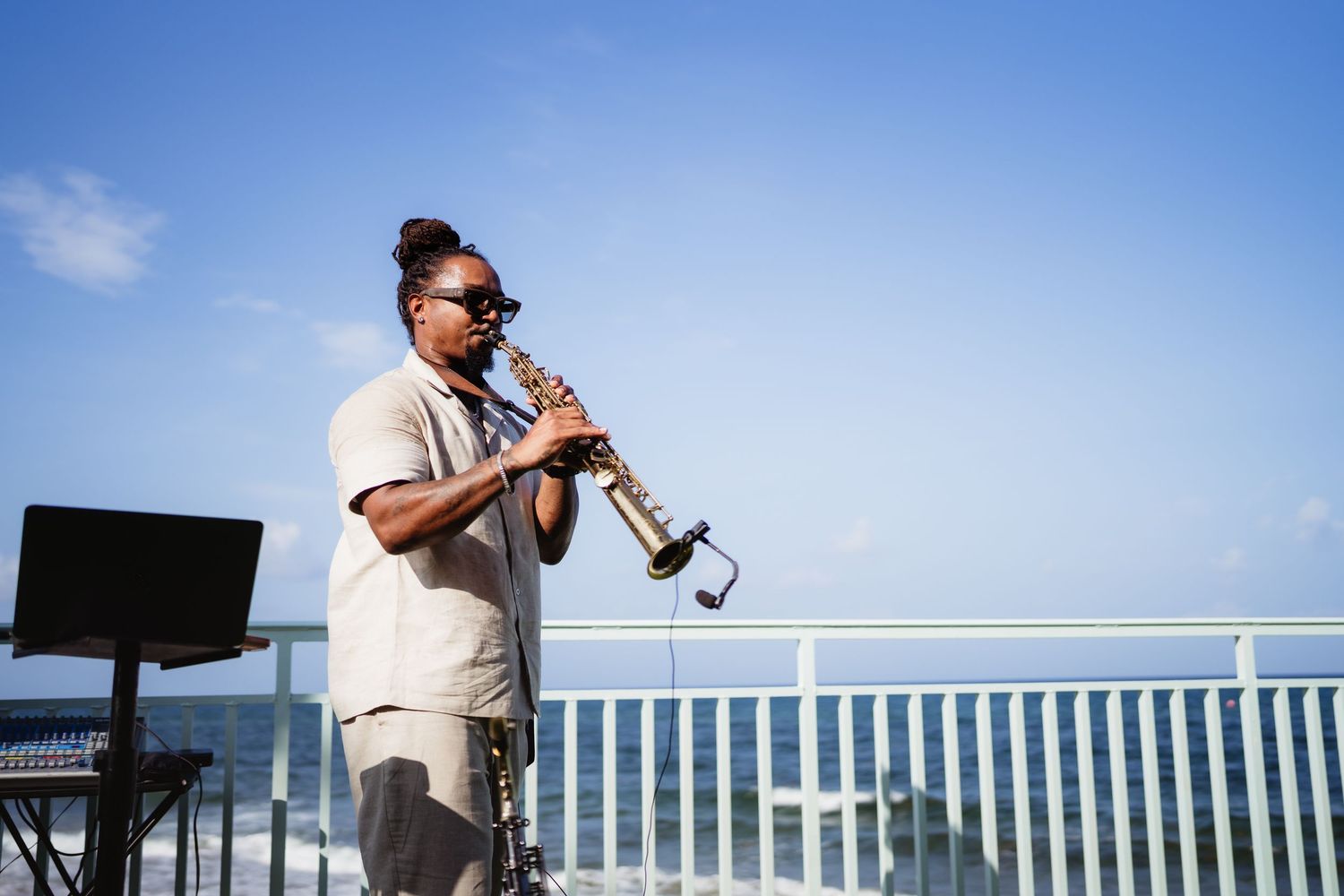 A musician plays saxophone on an outdoor rooftop stage with ocean views under a clear blue sky.