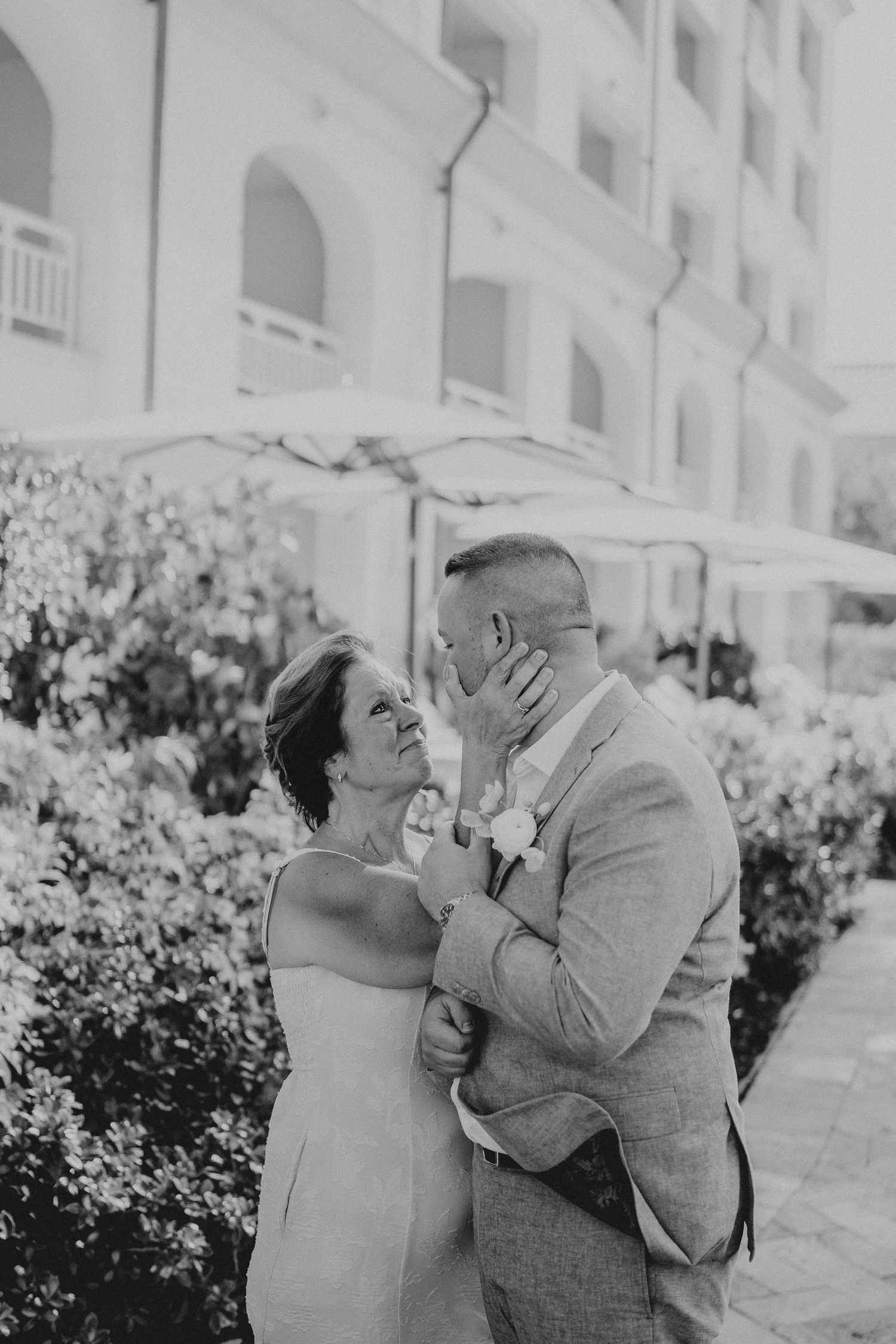 Black and white photo of a couple sharing a tender moment, the groom in a suit and bride in a dress, outdoors near lush greenery.