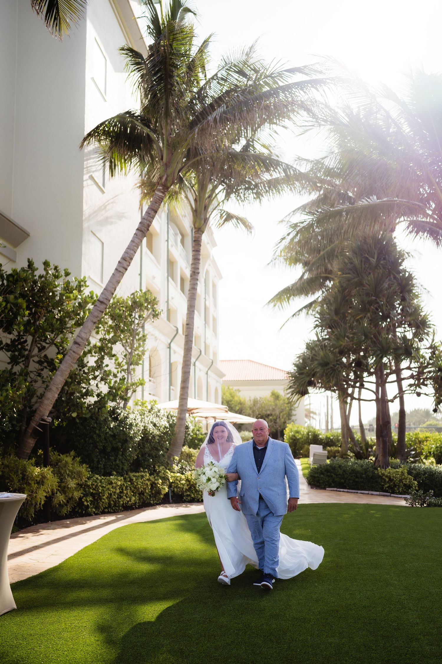 Bride and groom walking on lush green lawn beside palm trees and white resort building on sunny day.