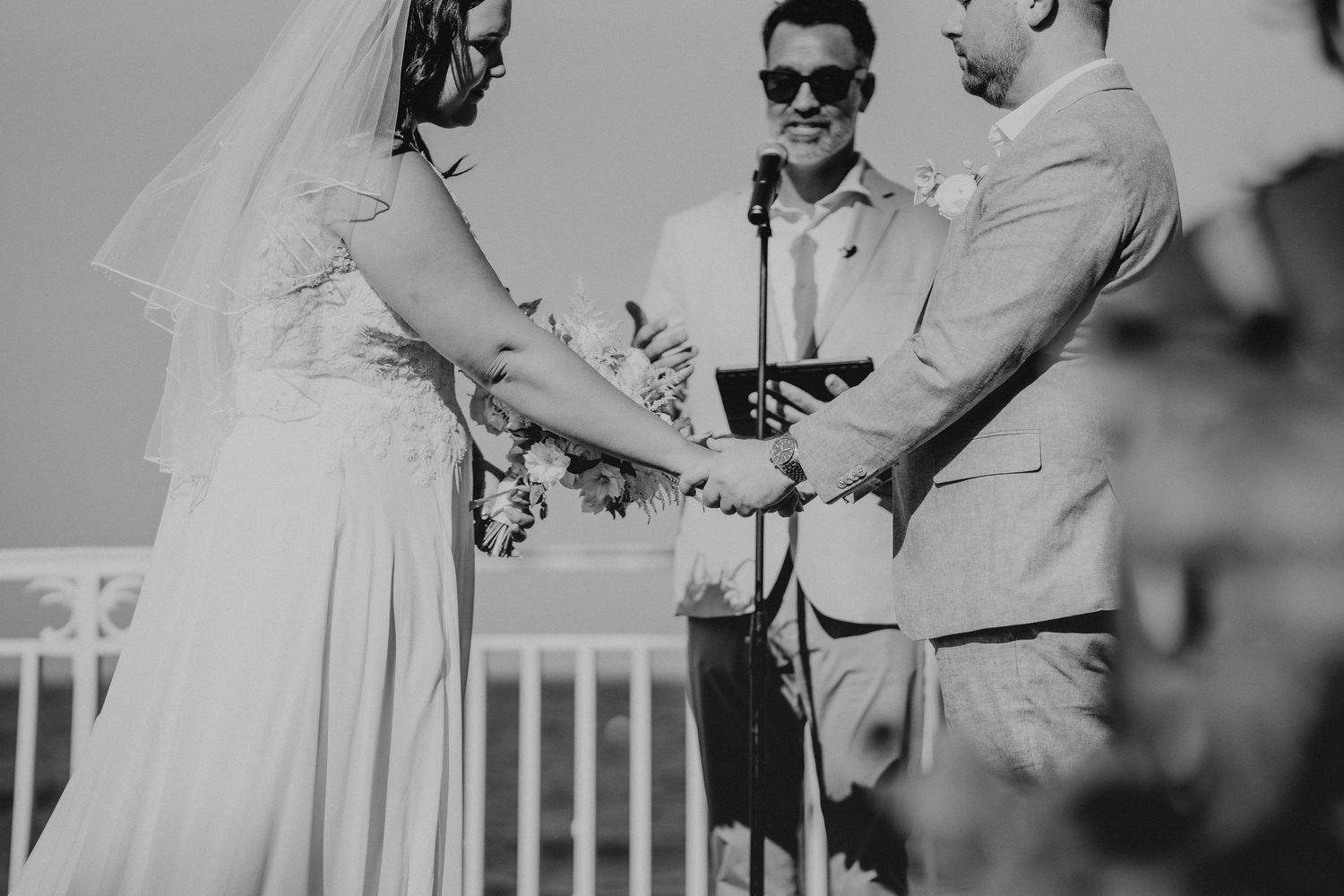 Black and white wedding ceremony photo of bride and groom exchanging vows with officiant wearing sunglasses outdoors.