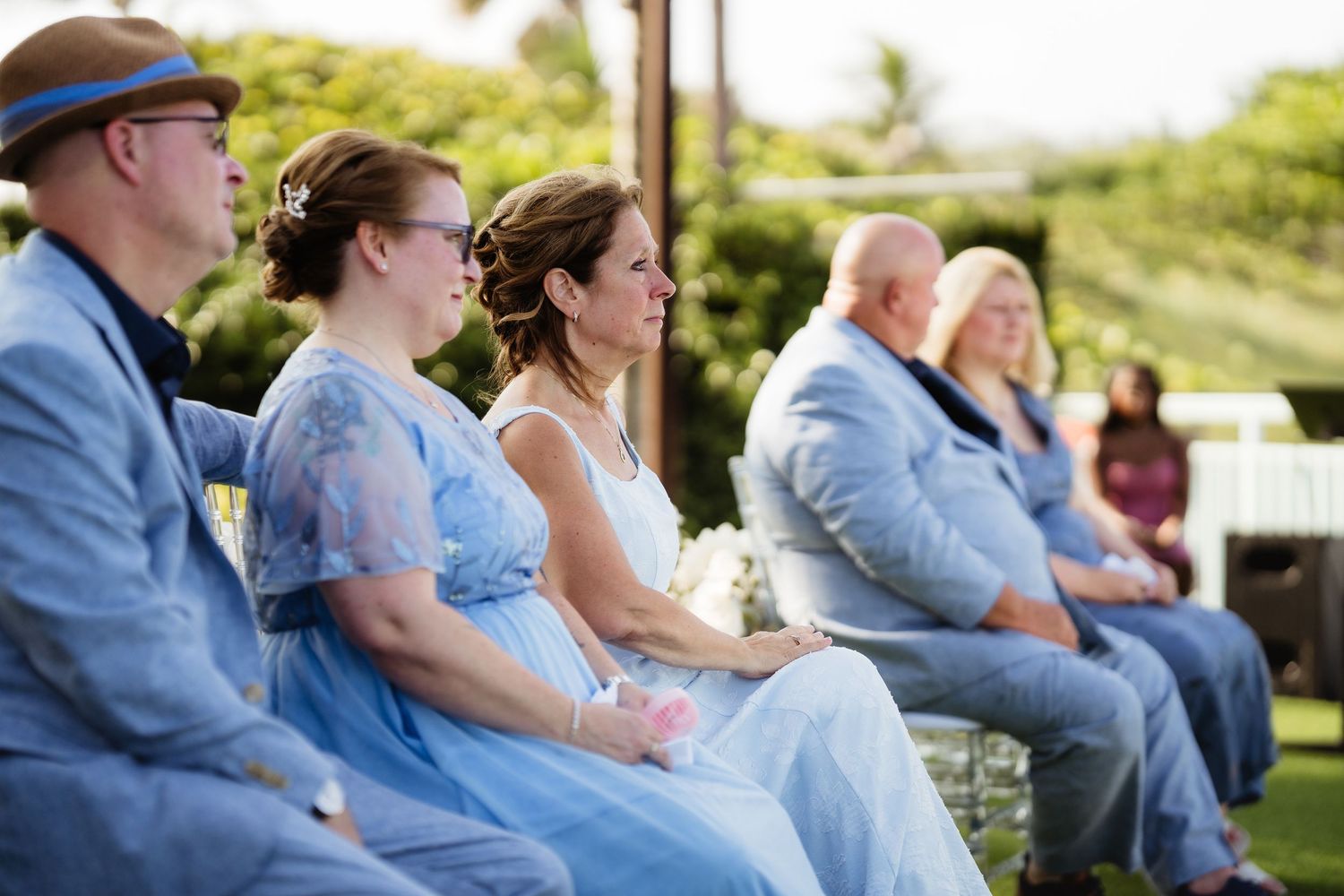 Guests in blue attire seated outdoors at a wedding ceremony on a sunny day.