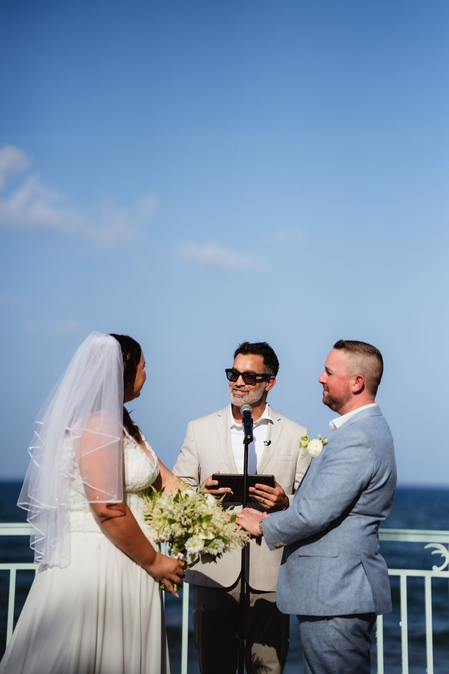 Bride and groom exchange vows with officiant at outdoor oceanfront wedding ceremony under clear blue sky.