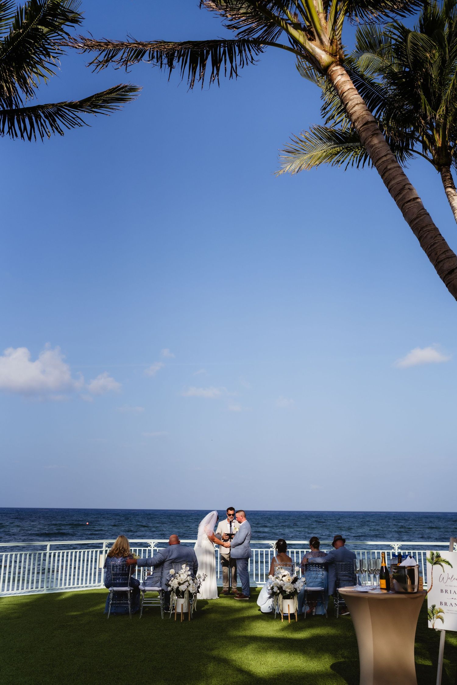 Outdoor beachside wedding ceremony on green lawn with palm trees, white arch, seated guests, and ocean backdrop under clear blue sky.