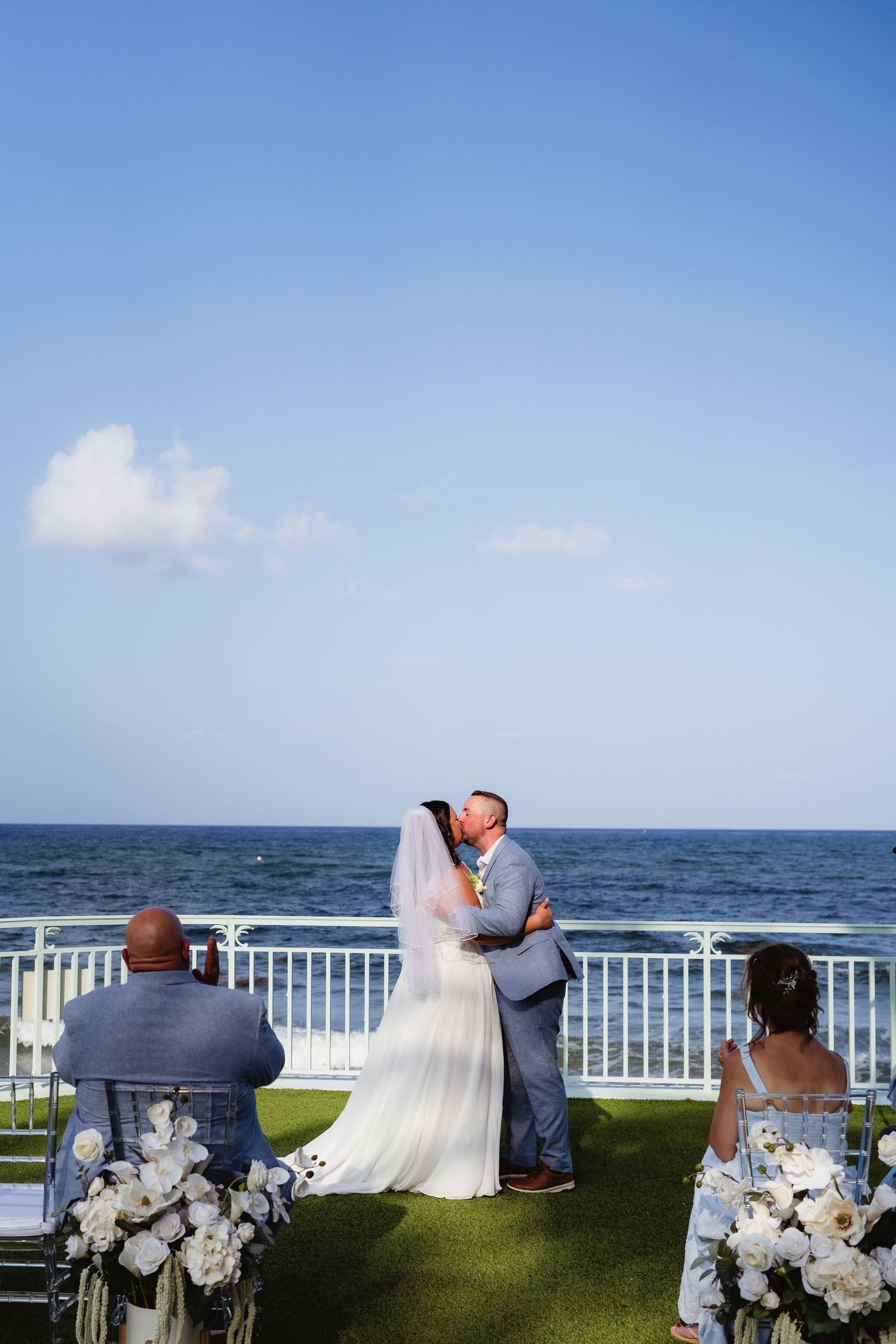 Bride and groom share first kiss at outdoor oceanfront wedding ceremony with white floral arrangements and blue sky backdrop.