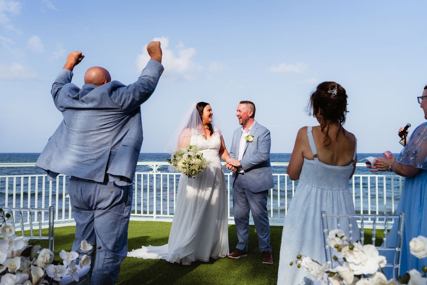 Bride and groom hold hands at outdoor oceanview wedding ceremony as officiant celebrates with raised arms.