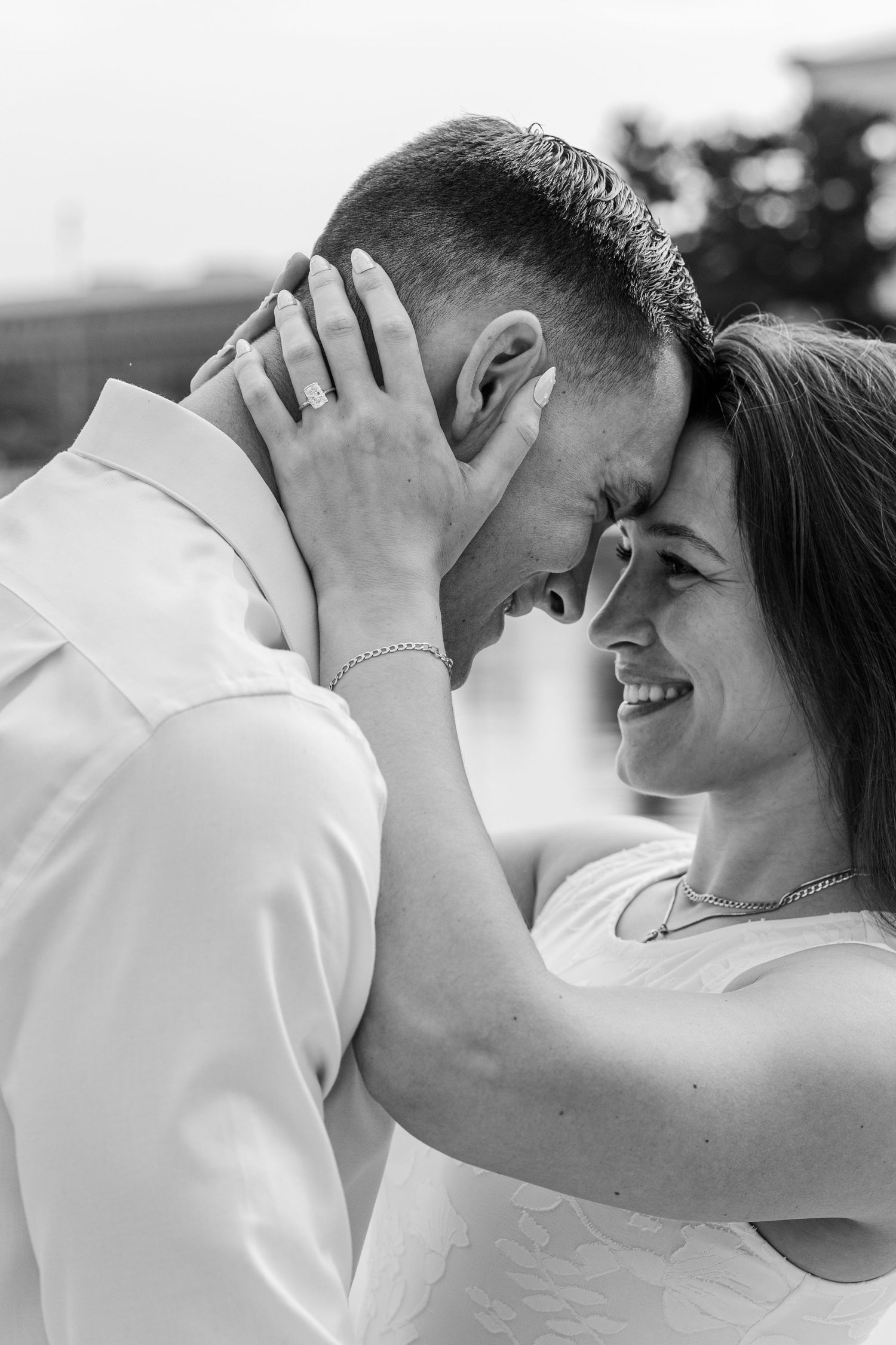Black and white photo of a smiling bride and groom touching foreheads, sharing a romantic moment outdoors.