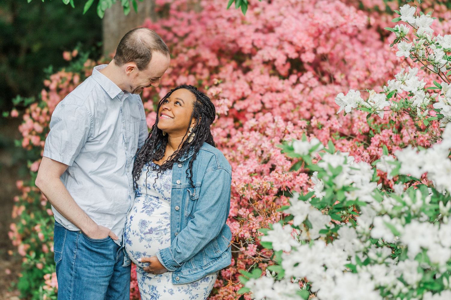 Expecting couple embracing among pink and white azalea blooms in a lush garden during a maternity photo session.