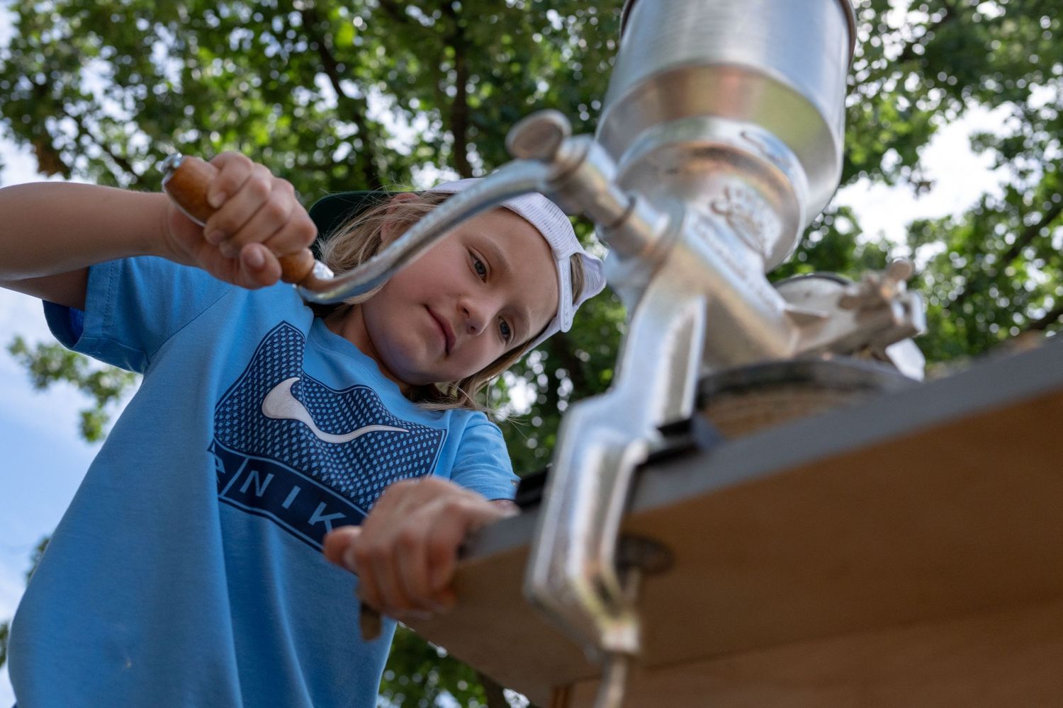 Young boy in blue Nike shirt using a hand pump water station outdoors on a sunny day.
