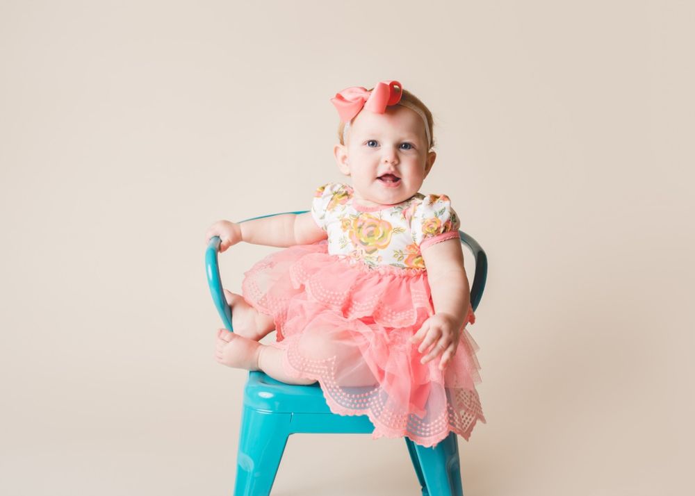 Smiling baby wearing a pink dress and bow while sitting on a small chair