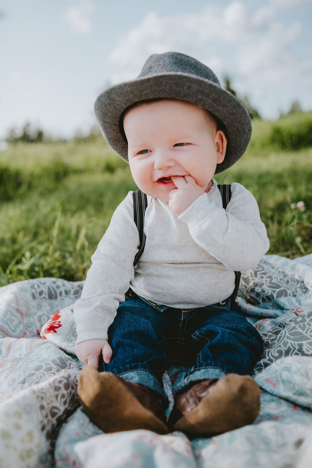 Smiling baby wearing a hat and sitting on a blanket in a grassy field during an outdoor portrait session