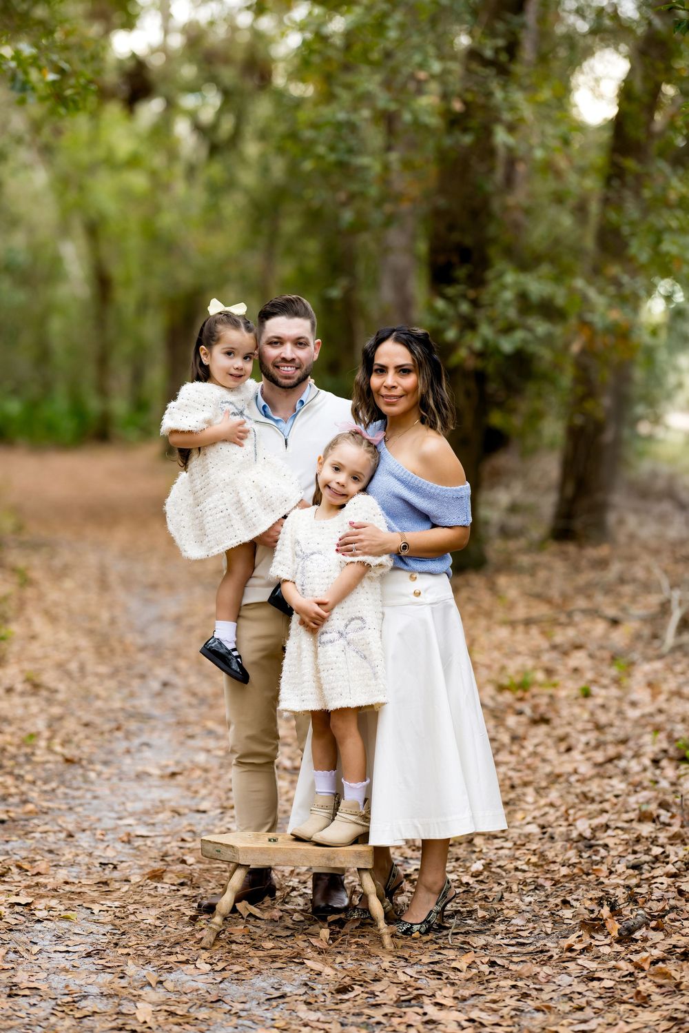 Family of four posing on a wooded path in autumn, parents holding two young daughters dressed in white outfits.