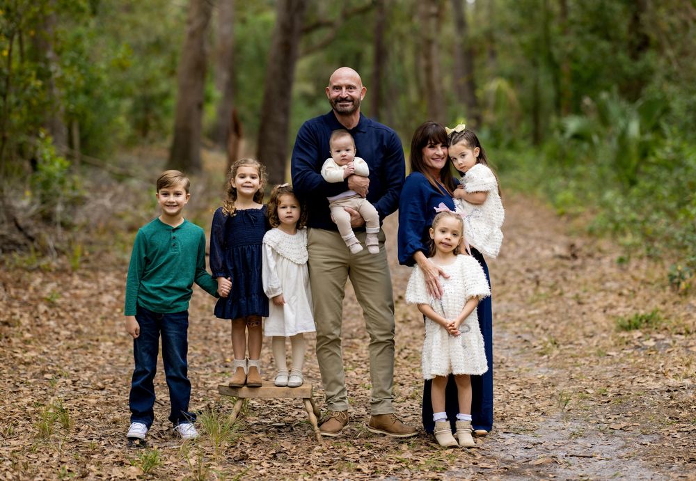 Family of eight posing on a forest path, parents with six young children smiling outdoors in coordinated outfits.