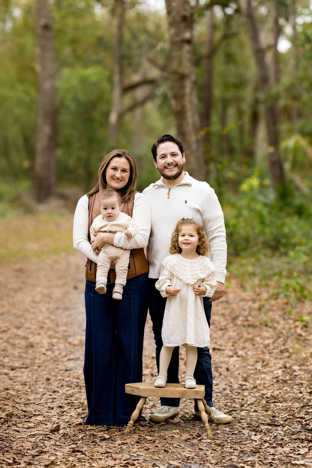 Family of four posing on a wooded trail, parents holding young children, dressed in neutral and navy tones.