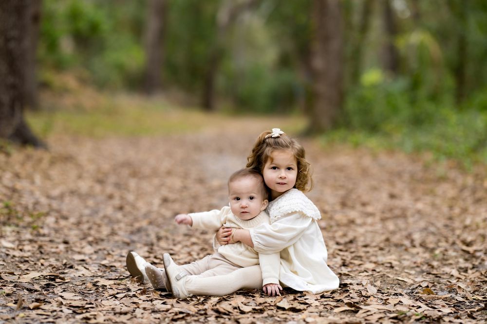 Two young sisters sitting on a leaf-covered forest path, older girl hugging baby sibling in white outfits.