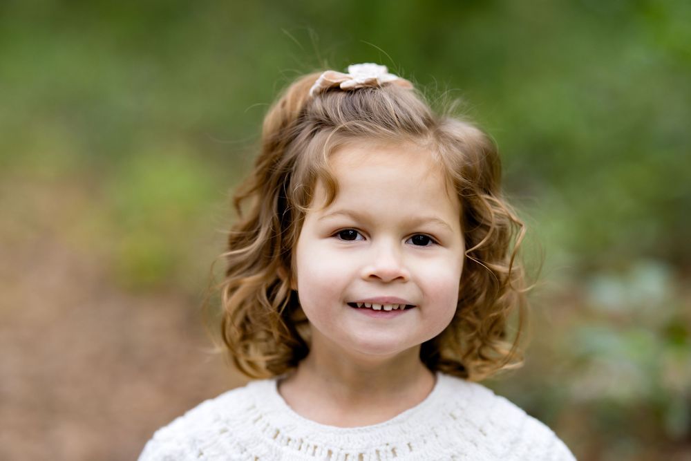 Smiling young girl with curly brown hair and a white sweater poses outdoors with a green nature background.