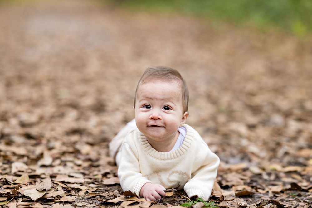 A smiling baby in a white sweater crawls on a bed of autumn leaves in a park.