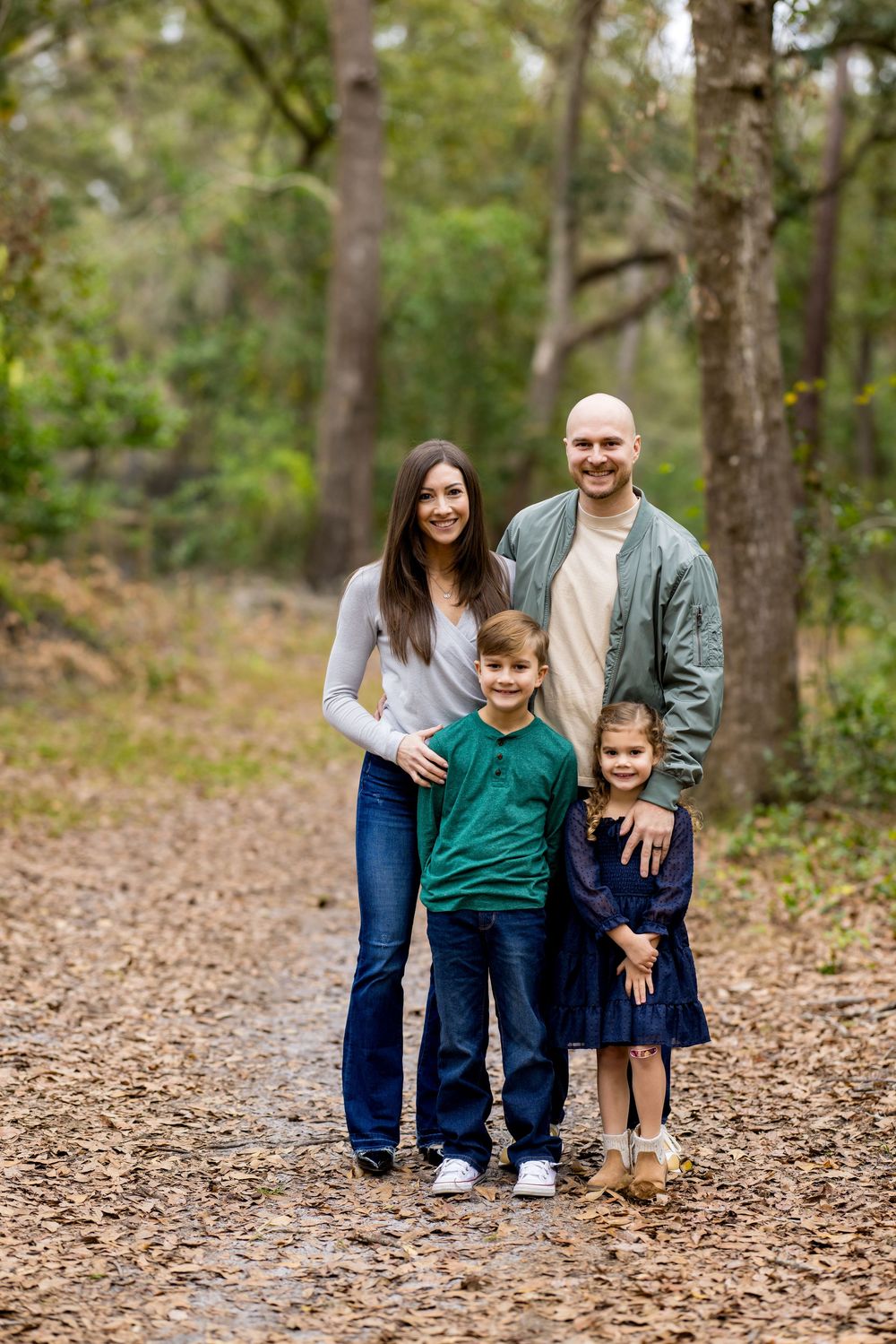Family of four posing on a leaf-covered forest path, parents standing behind two young children among tall trees.