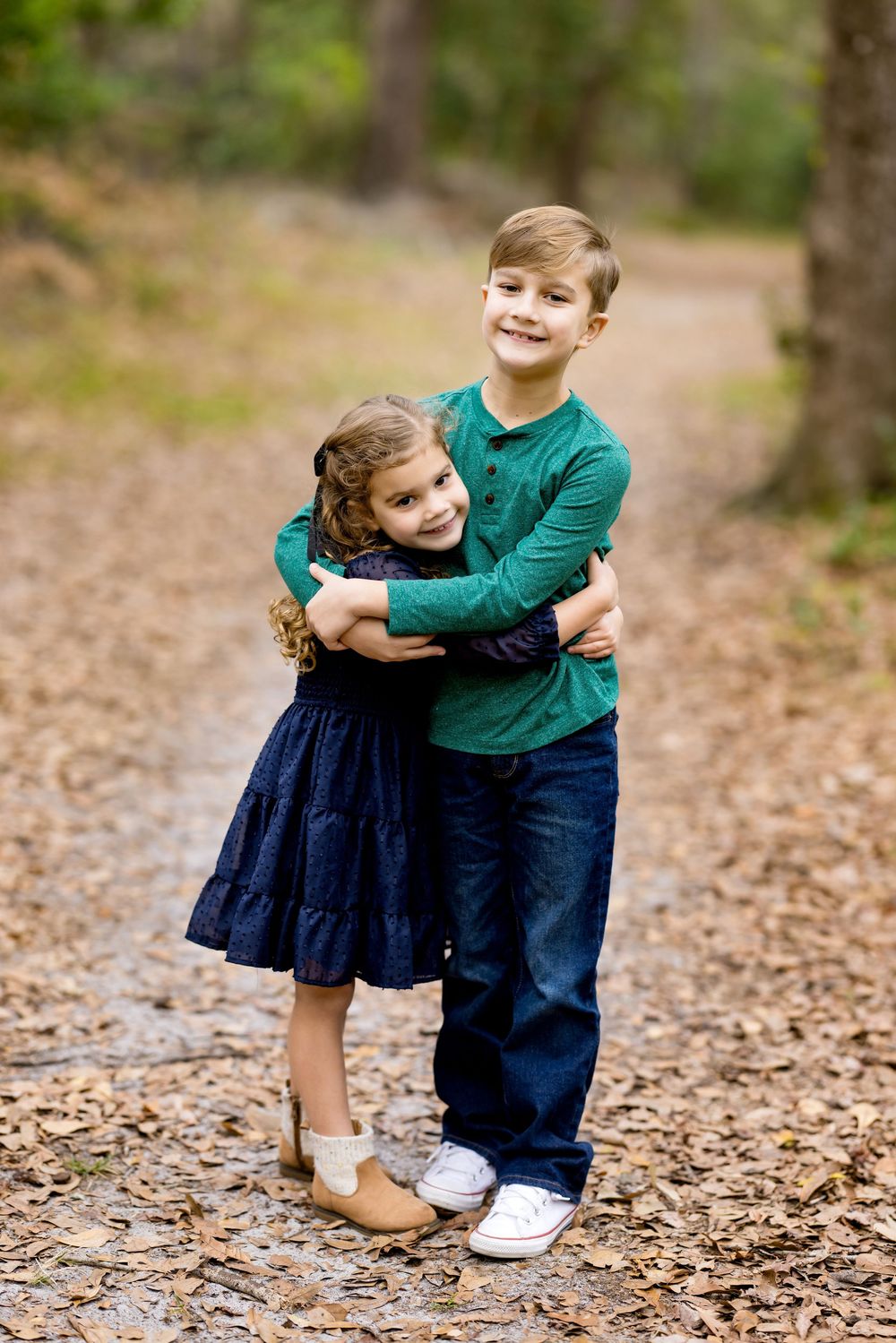 A smiling boy in a green shirt hugs a little girl in a navy dress on a leaf-covered forest path.