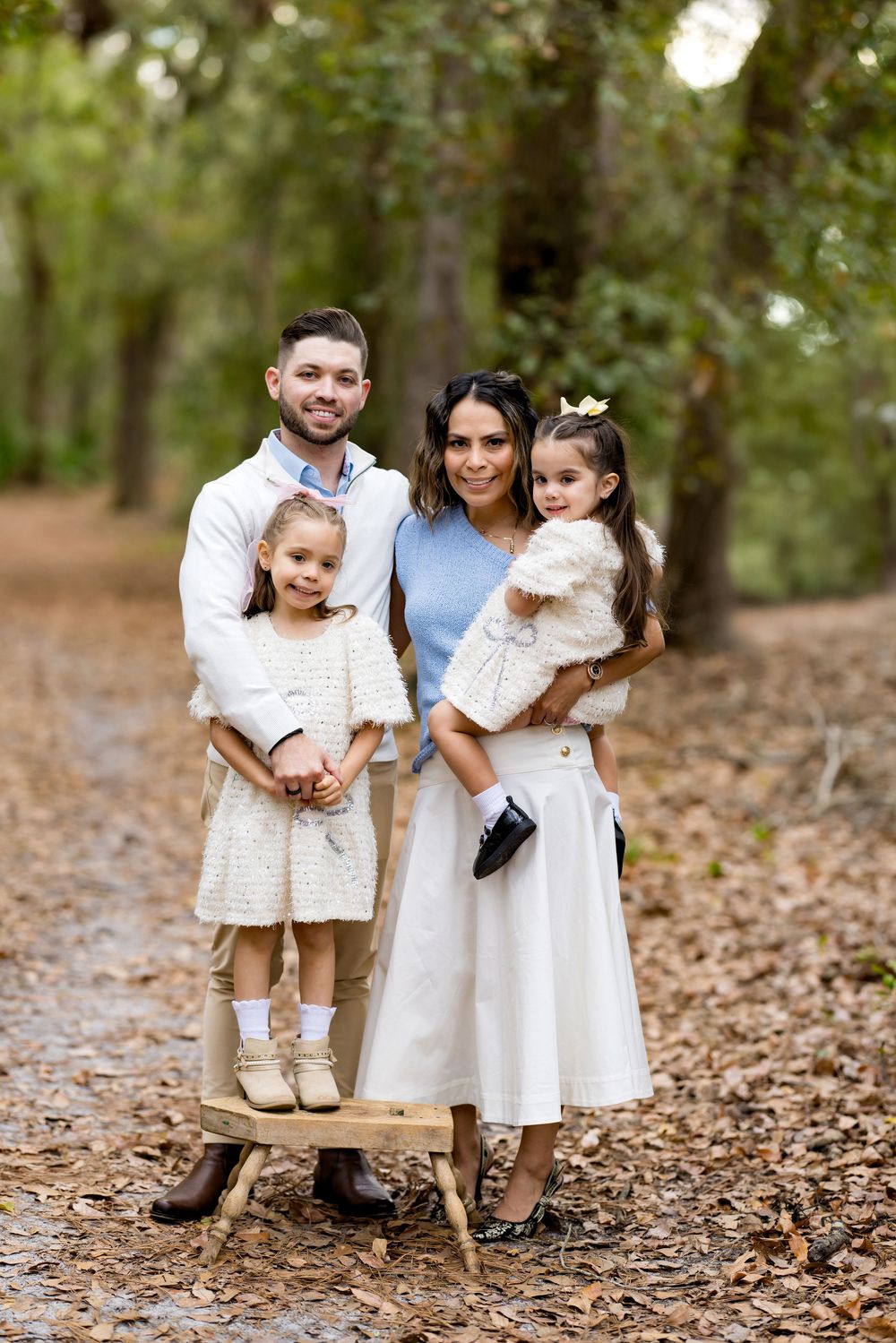 Family of four posing outdoors on a leaf-covered path surrounded by trees, parents holding two young girls in matching cream outfits.