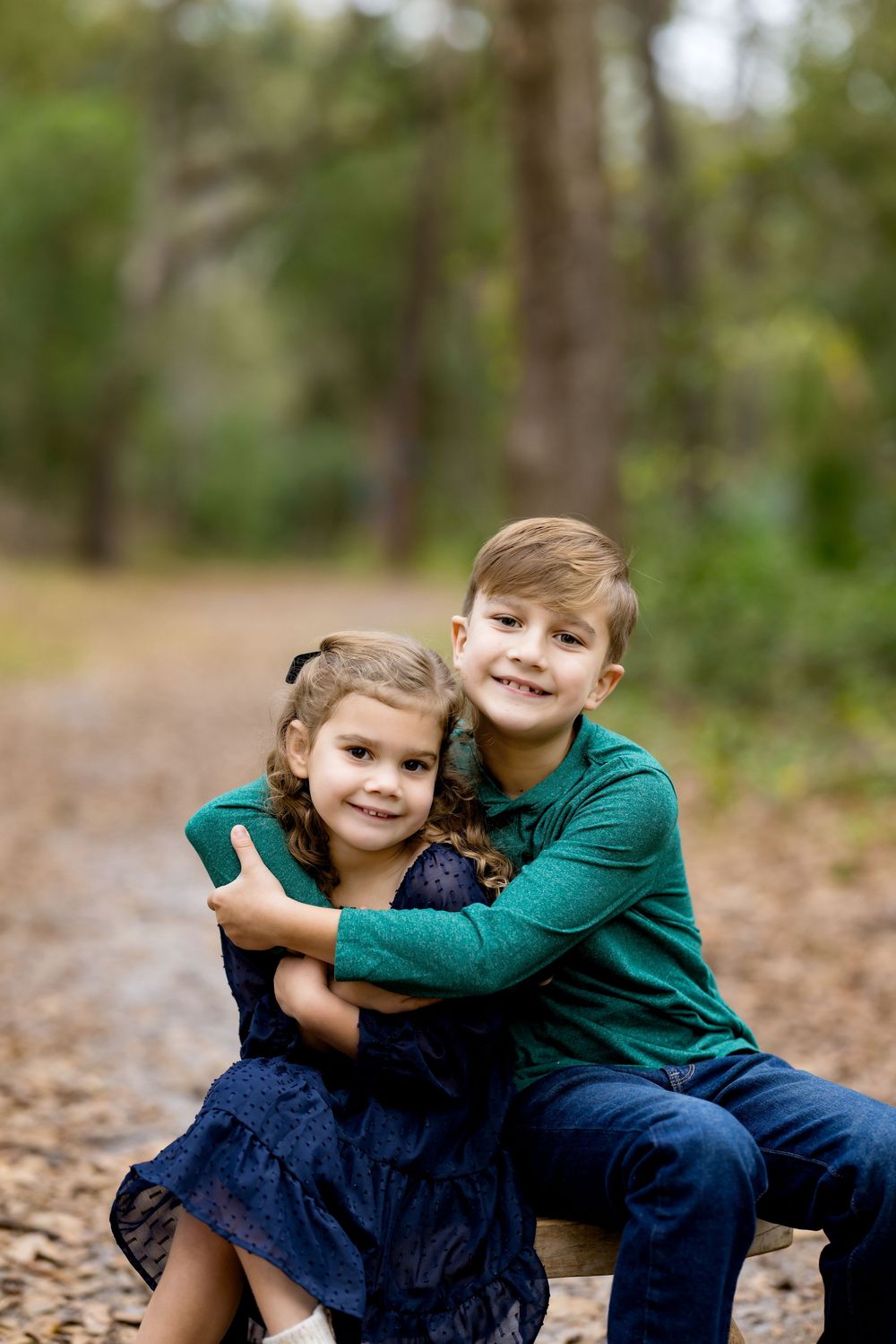 Two young siblings hugging outdoors on a forest path, boy in green shirt embracing girl in navy dress.