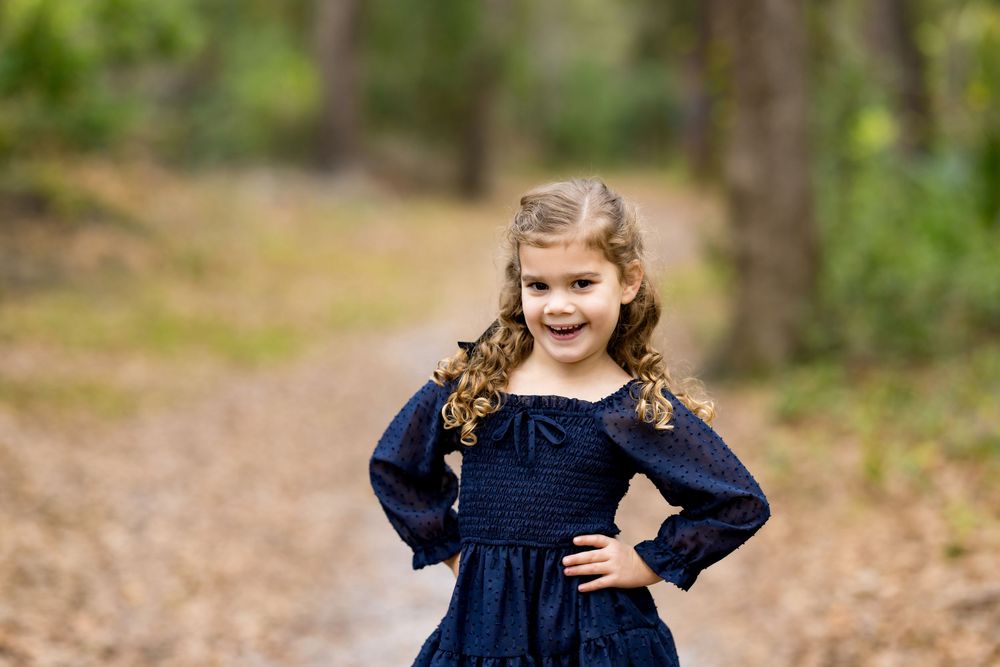 A young girl in a navy smocked dress poses confidently on a forest path with curly hair and hands on hips.