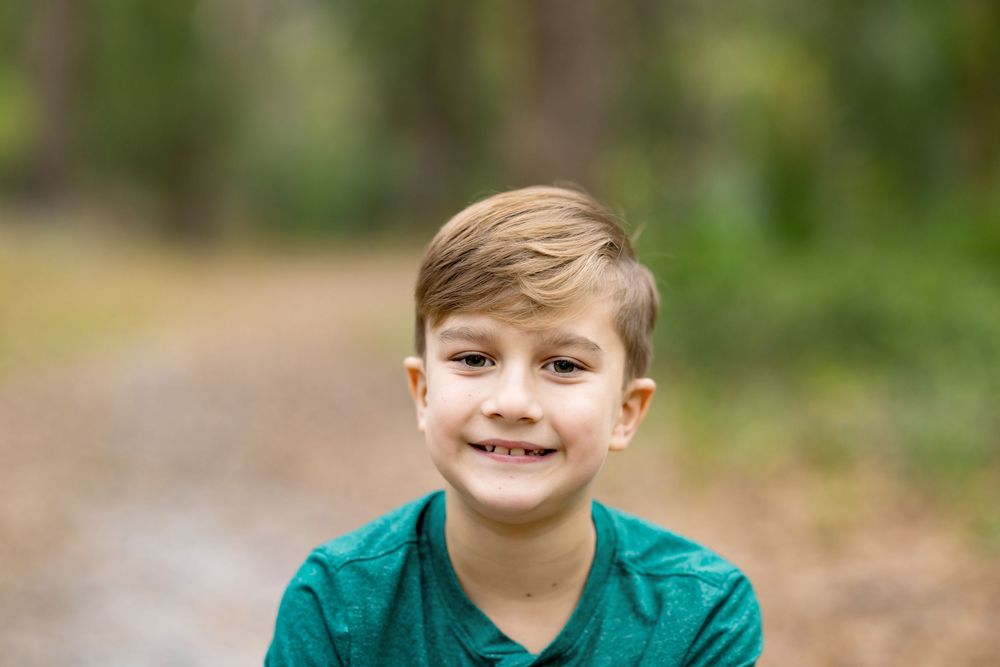 Smiling young boy with brown hair wearing a teal shirt, posing outdoors on a wooded path.