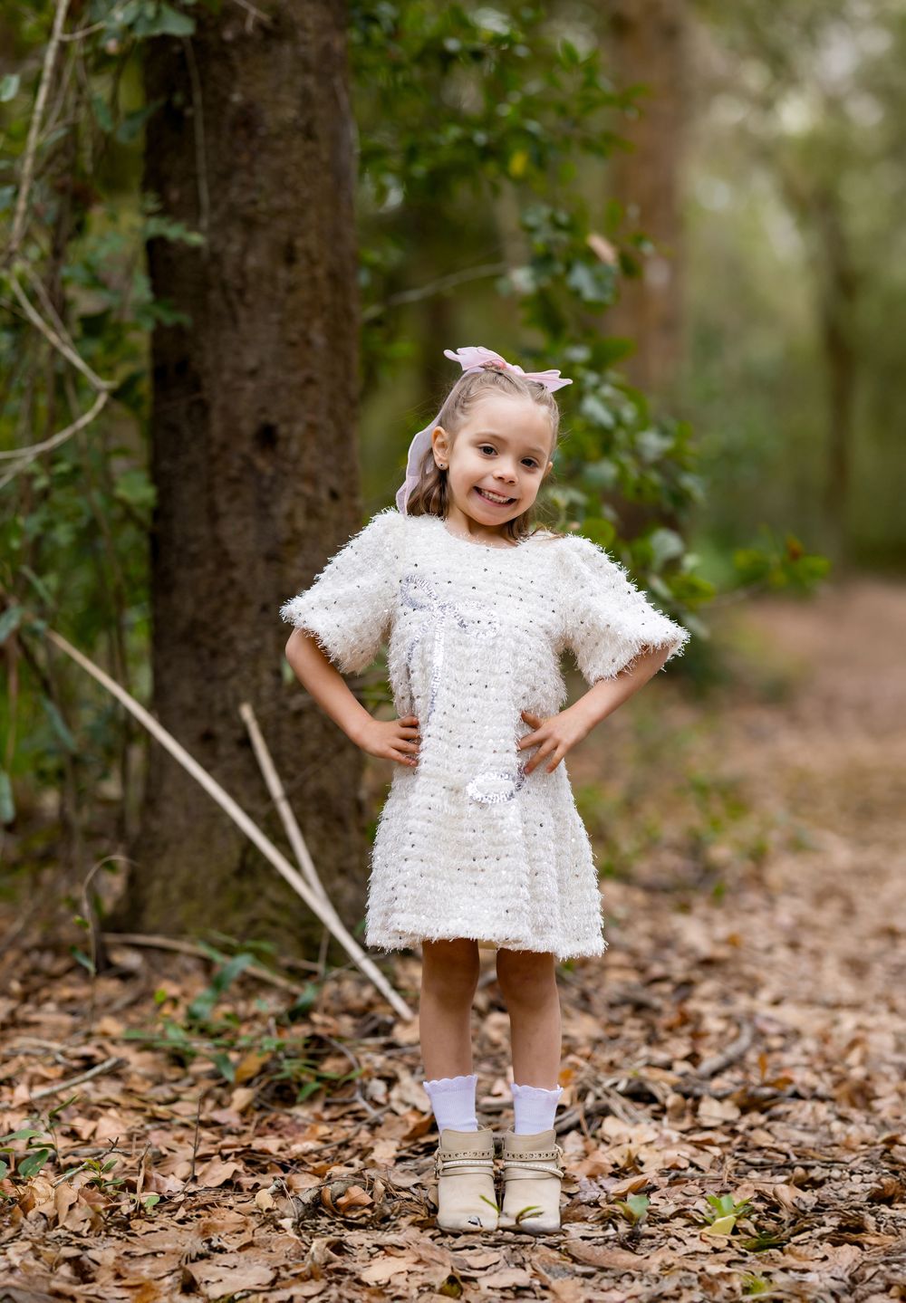 Young girl in white knit dress and boots poses with hands on hips on a forest path covered in autumn leaves.