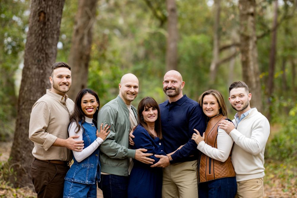 Seven people posing together outdoors in a wooded setting, smiling warmly in a family portrait photo.