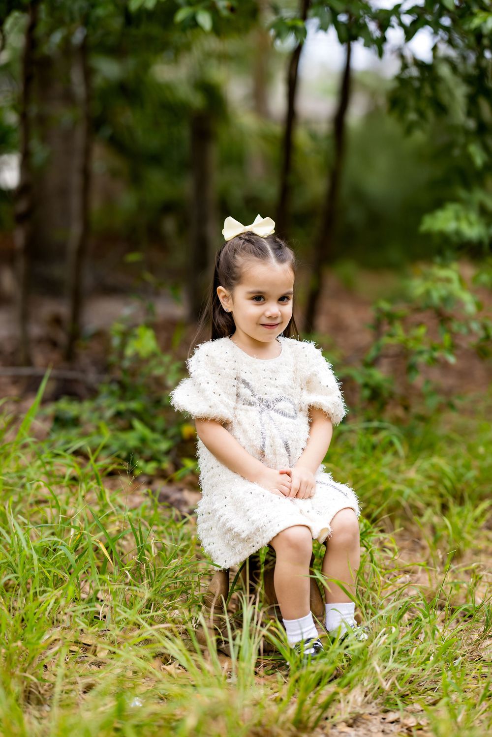 A young girl in a white fluffy dress with a bow sits outdoors in a lush green garden, smiling sweetly.