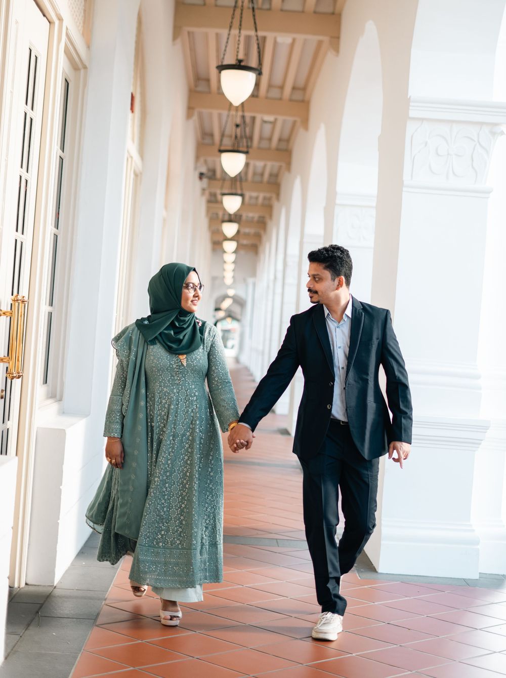 A Muslim couple walks hand-in-hand through an elegant colonial corridor, she in a teal hijab and embroidered dress, he in a dark suit.