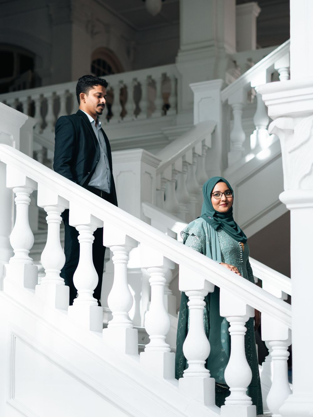 A man in a suit and a woman in a teal hijab descend elegant white stairs in a grand building.